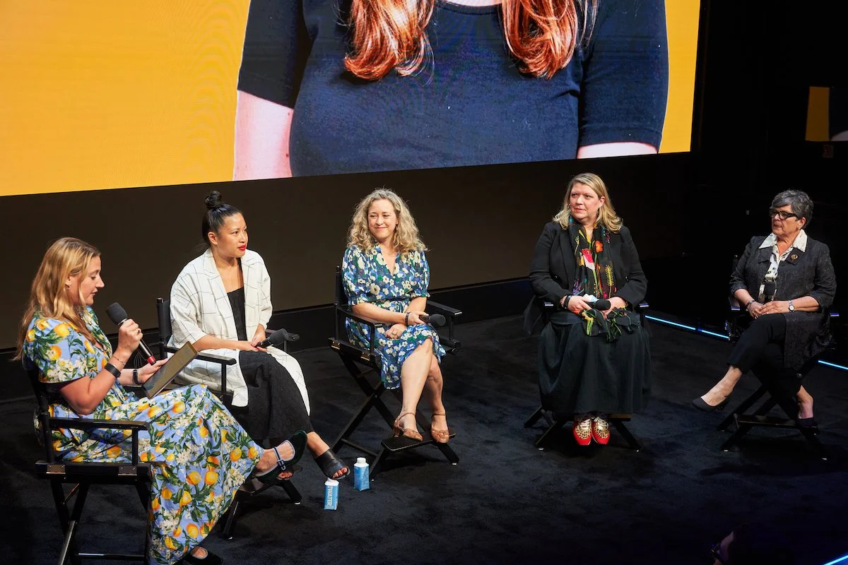 Panel of five women sitting on chairs on a stage during a discussion, with a large screen behind them displaying a close-up of one woman's torso in a black shirt.