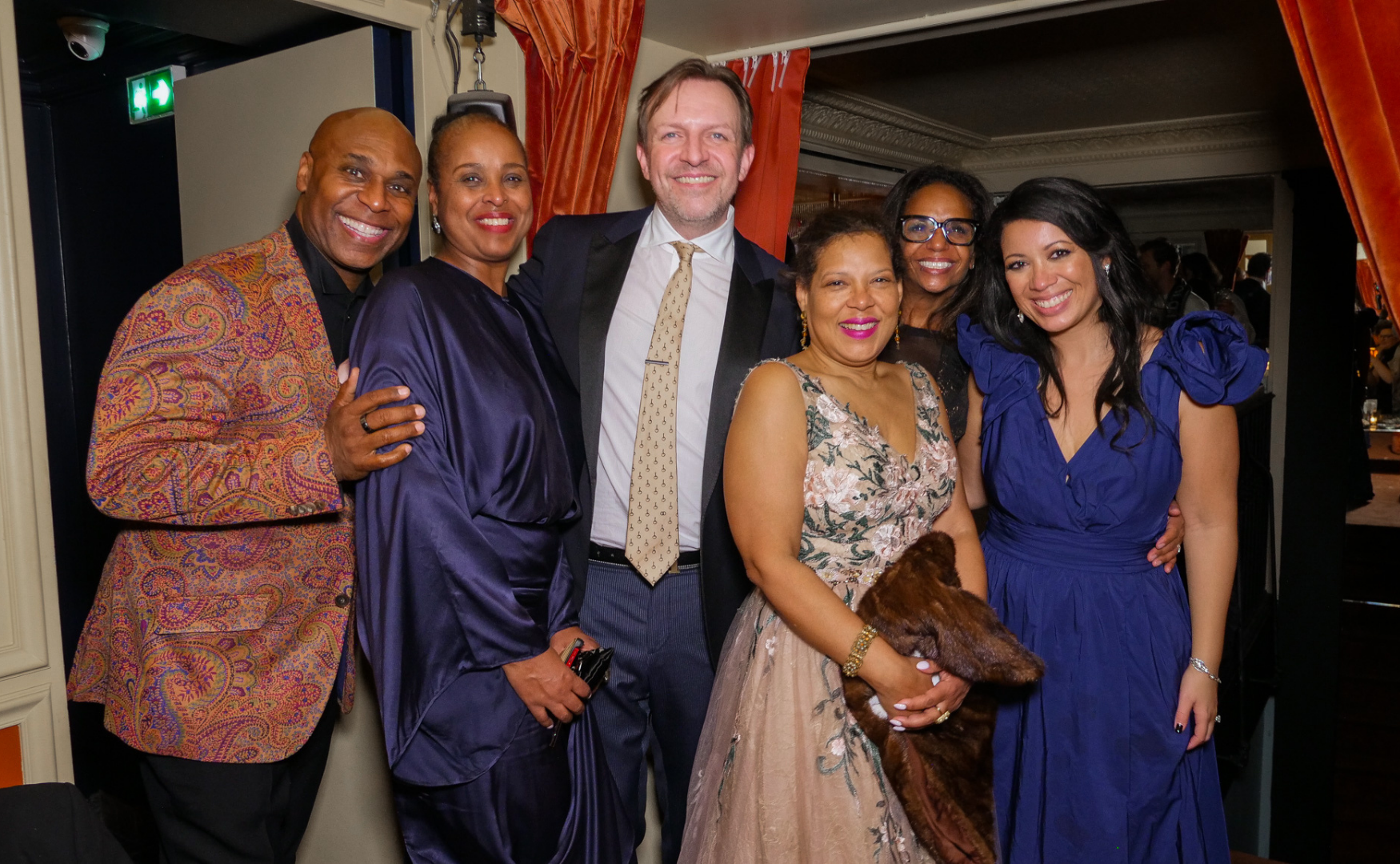 Group of six people posing together at a celebration, dressed in formal attire, smiling, indoors with orange curtains and a dimly lit background.