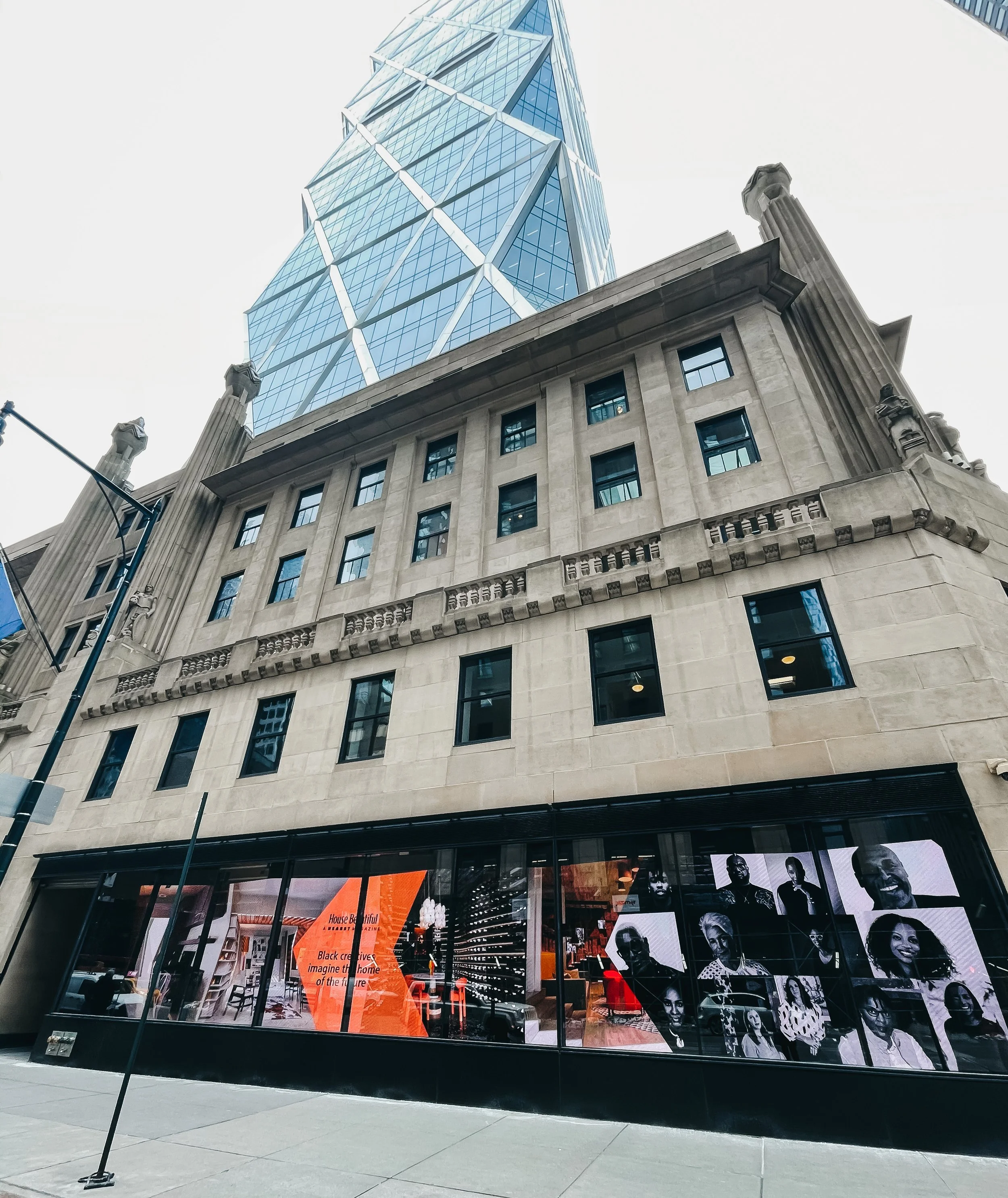A city street view showing a modern skyscraper with glass panels above an older, classical-style building with large windows and decorative columns. The ground floor has a storefront with portraits in black and white.