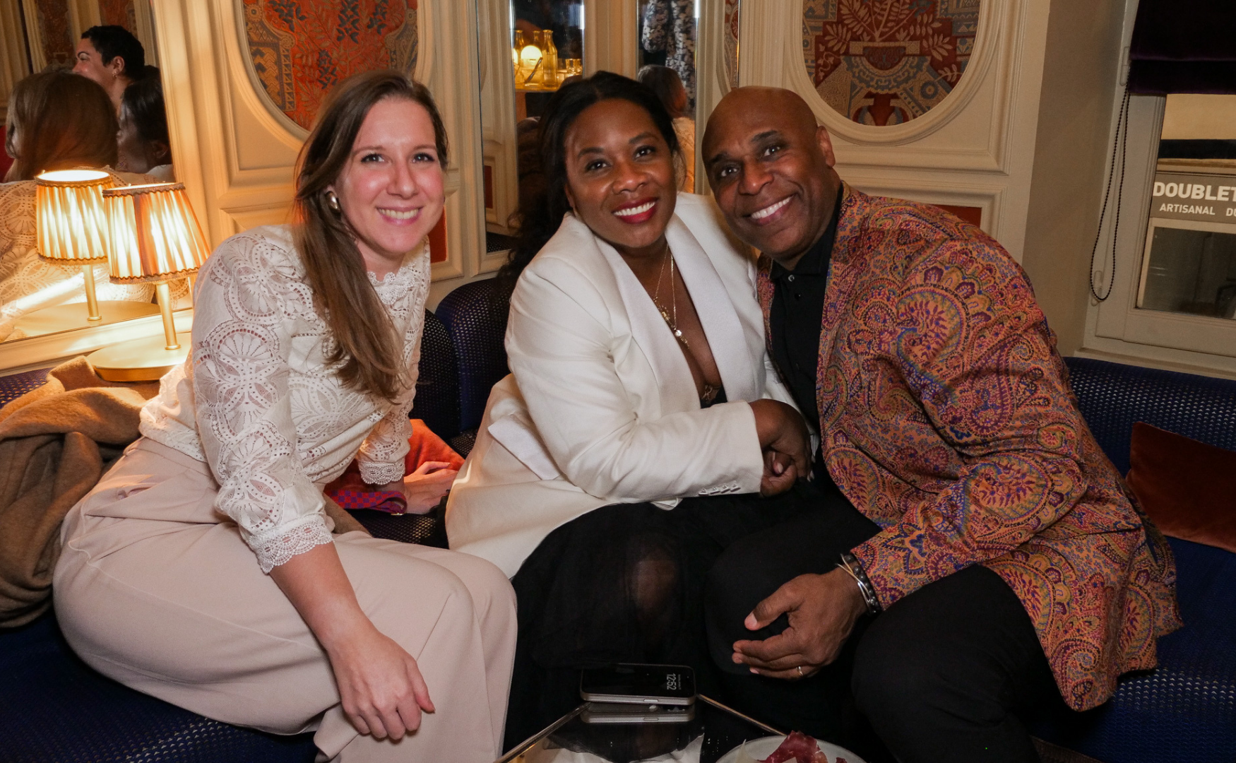Three people smiling and sitting close together on a sofa in a cozy, warmly lit restaurant or lounge. The woman on the left has long light brown hair, is wearing a white eyelet blouse and cream-colored pants. The woman in the middle has dark hair, is