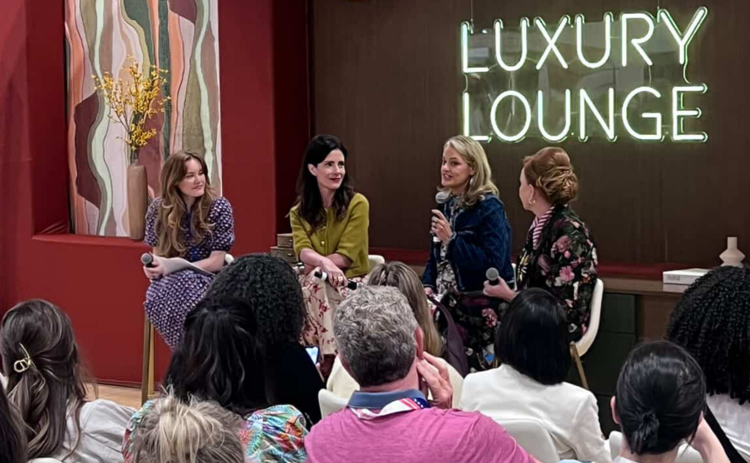 Four women seated on a stage in front of an audience during a panel discussion at a venue with a large neon sign reading 'LUXURY LOUNGE'. One woman is holding a microphone and speaking, while the others listen. The stage has decorative wall art and a