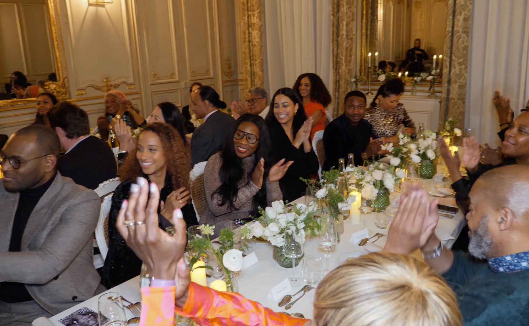 People sitting at a decorated banquet table, clapping and smiling during an elegant event in a luxurious room with ornate gold details and floral arrangements.