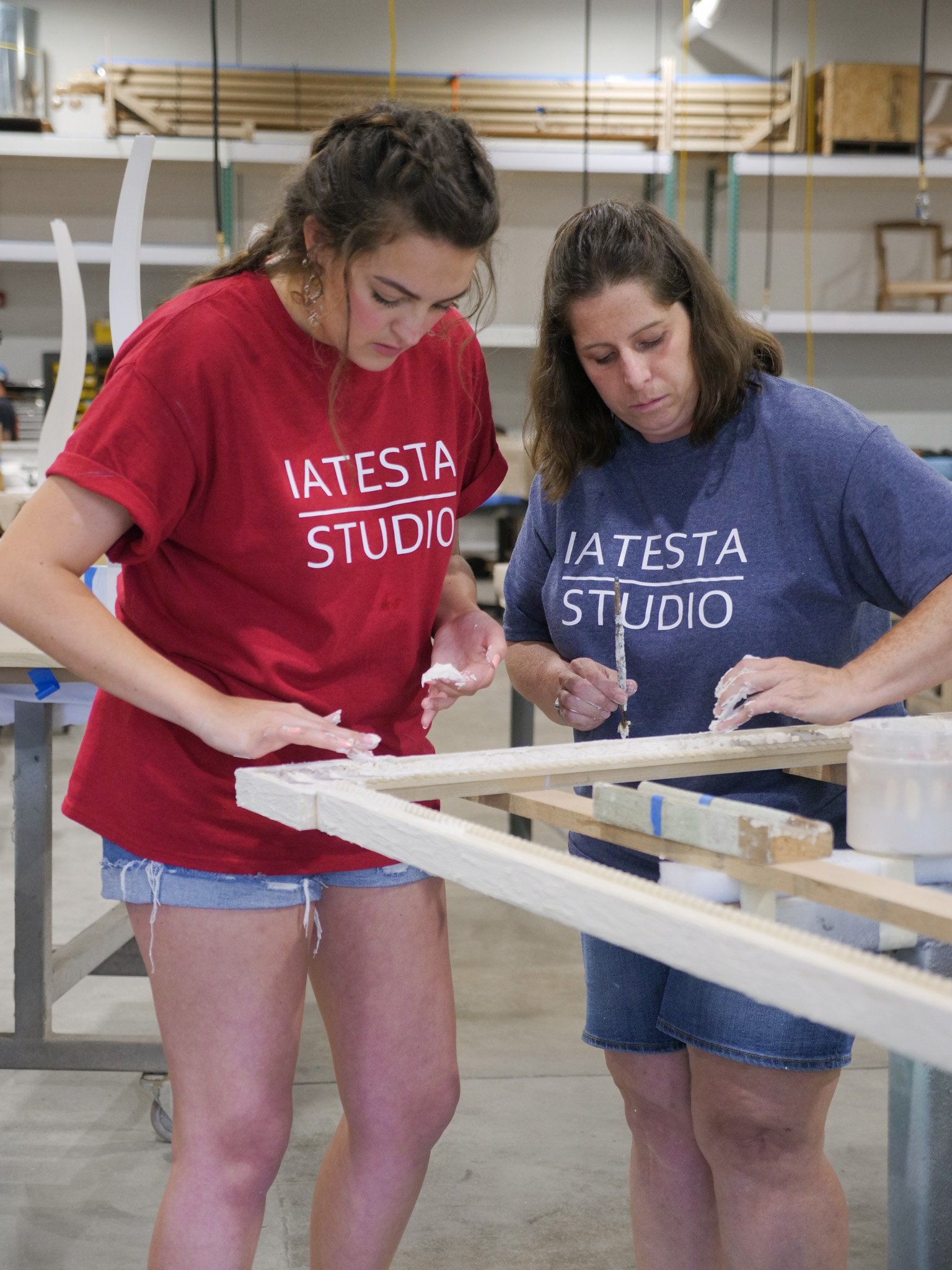 Two women working on a woodworking project in a workshop, wearing T-shirts with the text 'I A T E S T A STUDIO', one in red and one in blue, focused on the project.