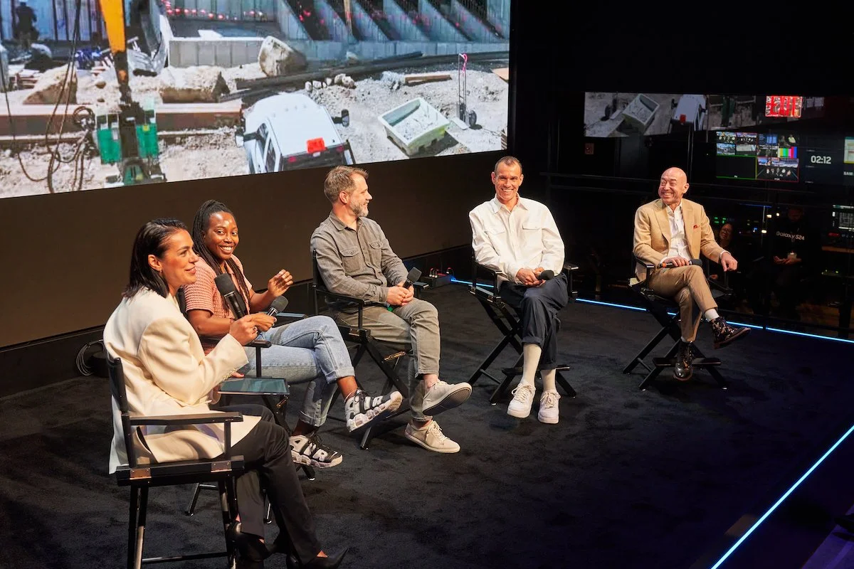 Five people sitting on chairs on stage, participating in a panel discussion. Behind them, a large screen displays an outdoor scene with rocks, construction materials, and a vehicle.