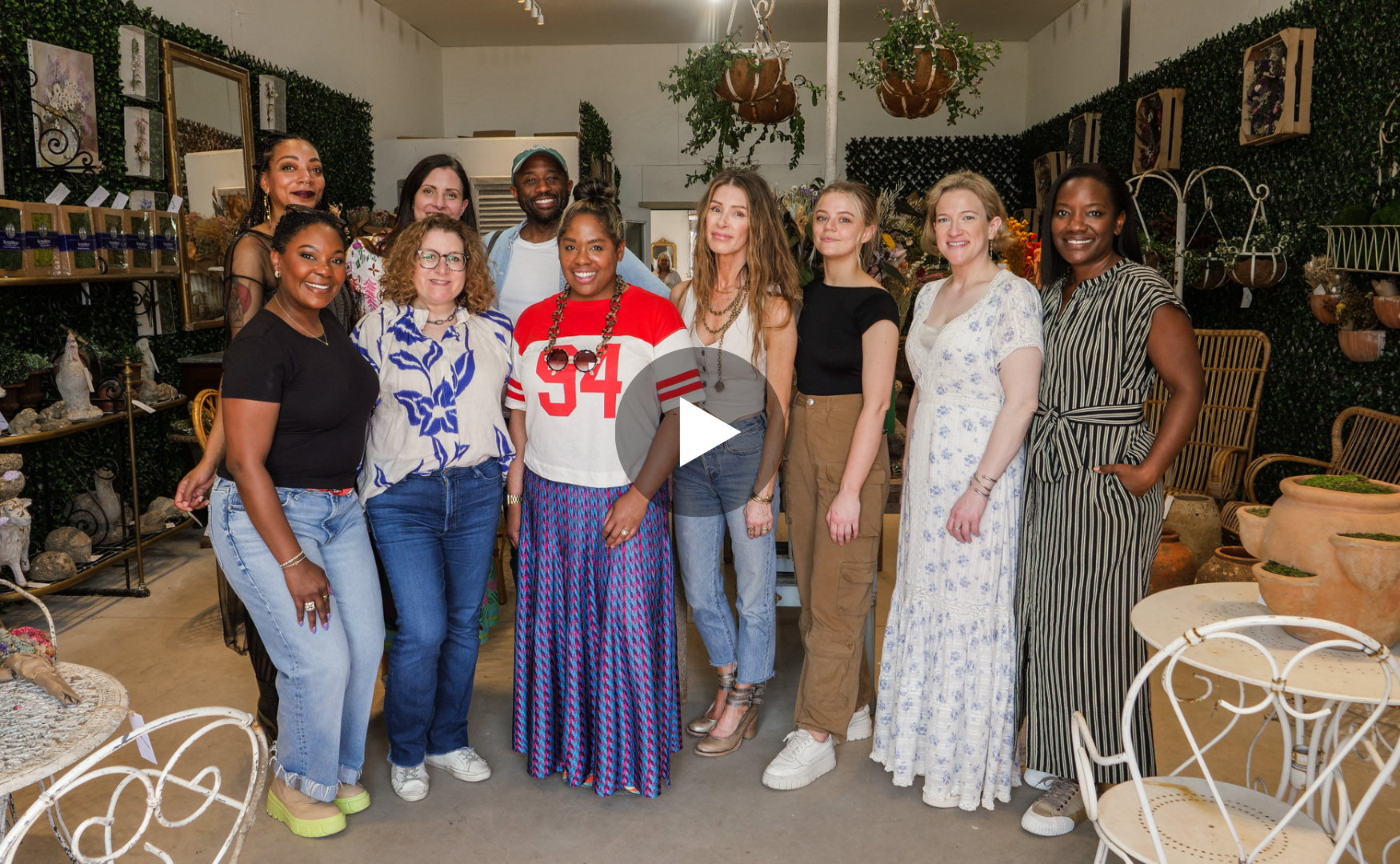 A group of twelve diverse people standing together in a cozy indoor space decorated with plants, artworks, and rustic furniture. They are smiling at the camera.
