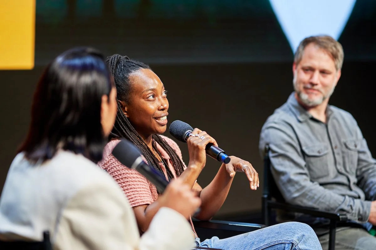 Three people sitting on stage participating in a panel discussion, one woman speaking into a microphone while the others listen.