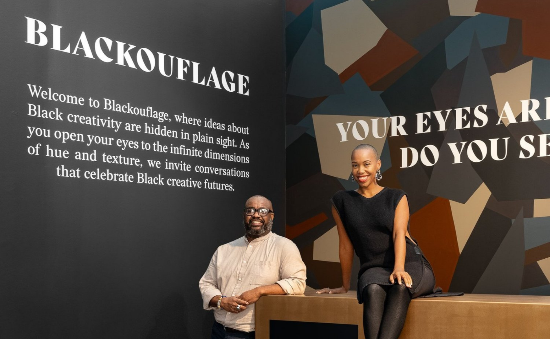 A man and woman posing in front of an art display at Blackuflage. The display has text about celebrating Black creativity and a colorful geometric background. The woman is sitting on a ledge, smiling, wearing a black top and earrings. The man is stan
