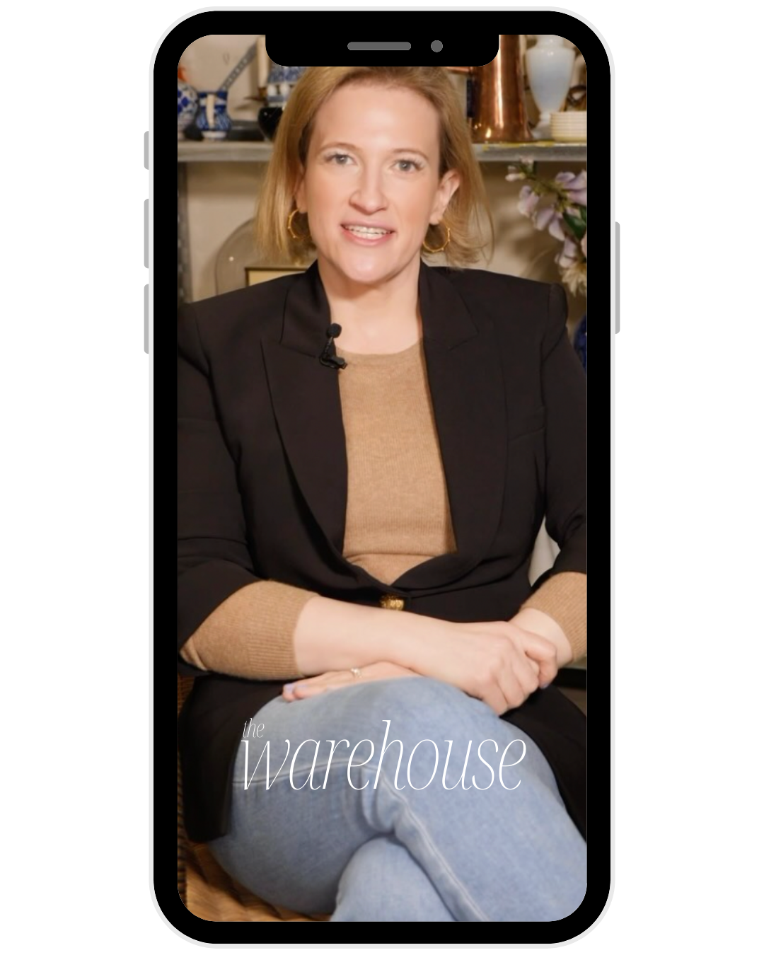 A woman with shoulder-length blonde hair, wearing a black blazer and a tan top, sitting with her hands on her lap, speaking in front of a shelving unit with decorative items. Text overlay reads 'the warehouse'.