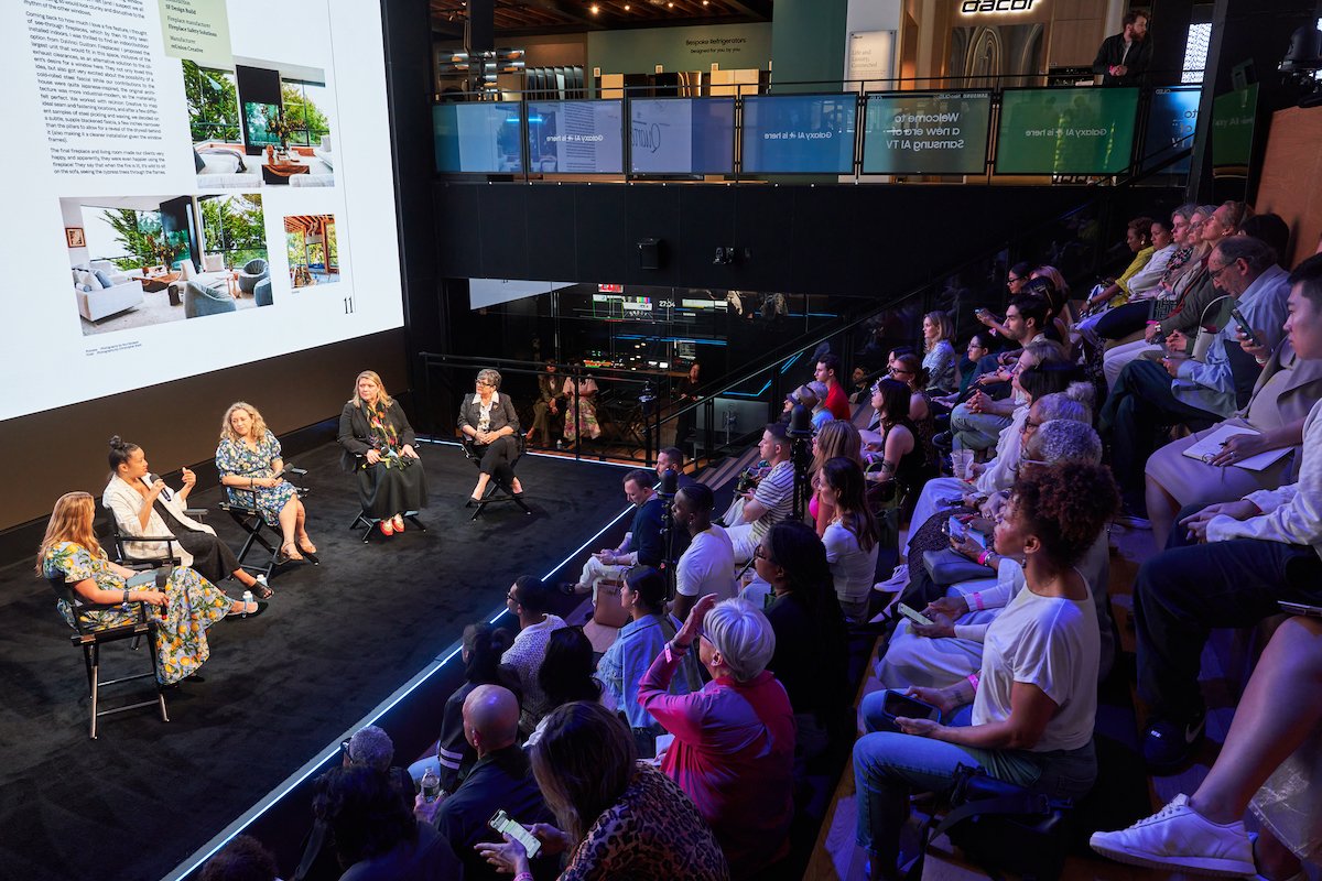 A panel discussion taking place on a stage with four women seated and speaking into microphones, in front of a large screen displaying presentation slides, with an audience seated in tiered seating watching attentively.