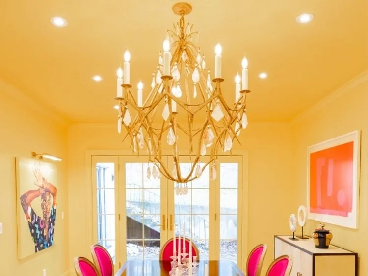 A dining room with a chandelier, pink chairs, a table with candles, a colorful painting, and a glass door.