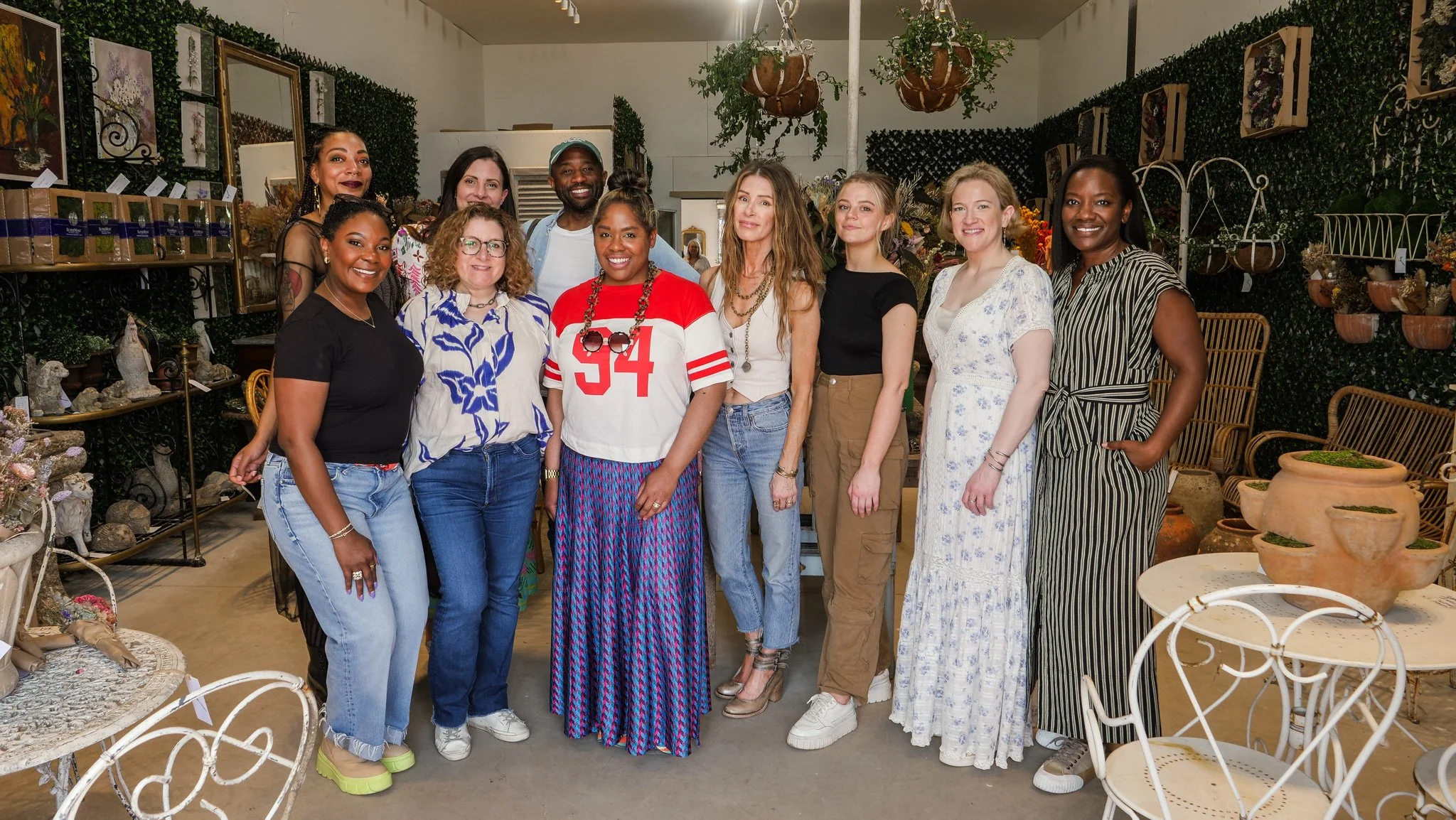 Group of ten women posing together inside a decor shop surrounded by plants, artwork, and decorative items.