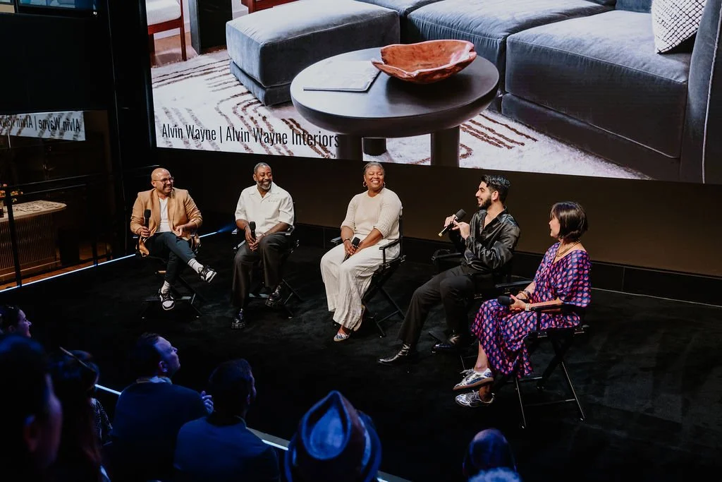 A panel discussion with five people seated on stage, three men and two women, engaging with an audience. A large screen behind them displays an interior design scene with a coffee table and couch.