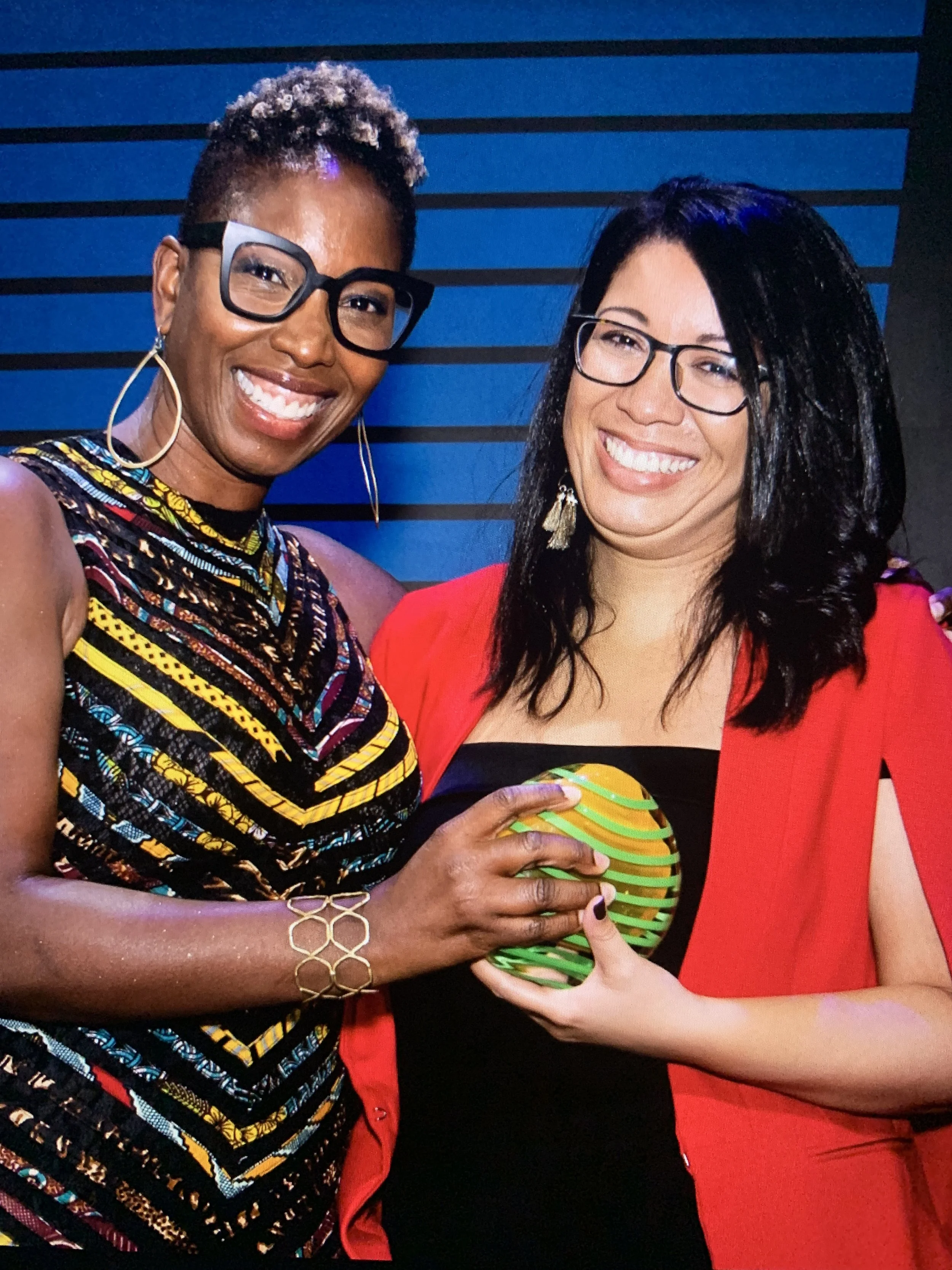 Two women smiling and holding a colorful object together, standing in front of a blue and black striped background.