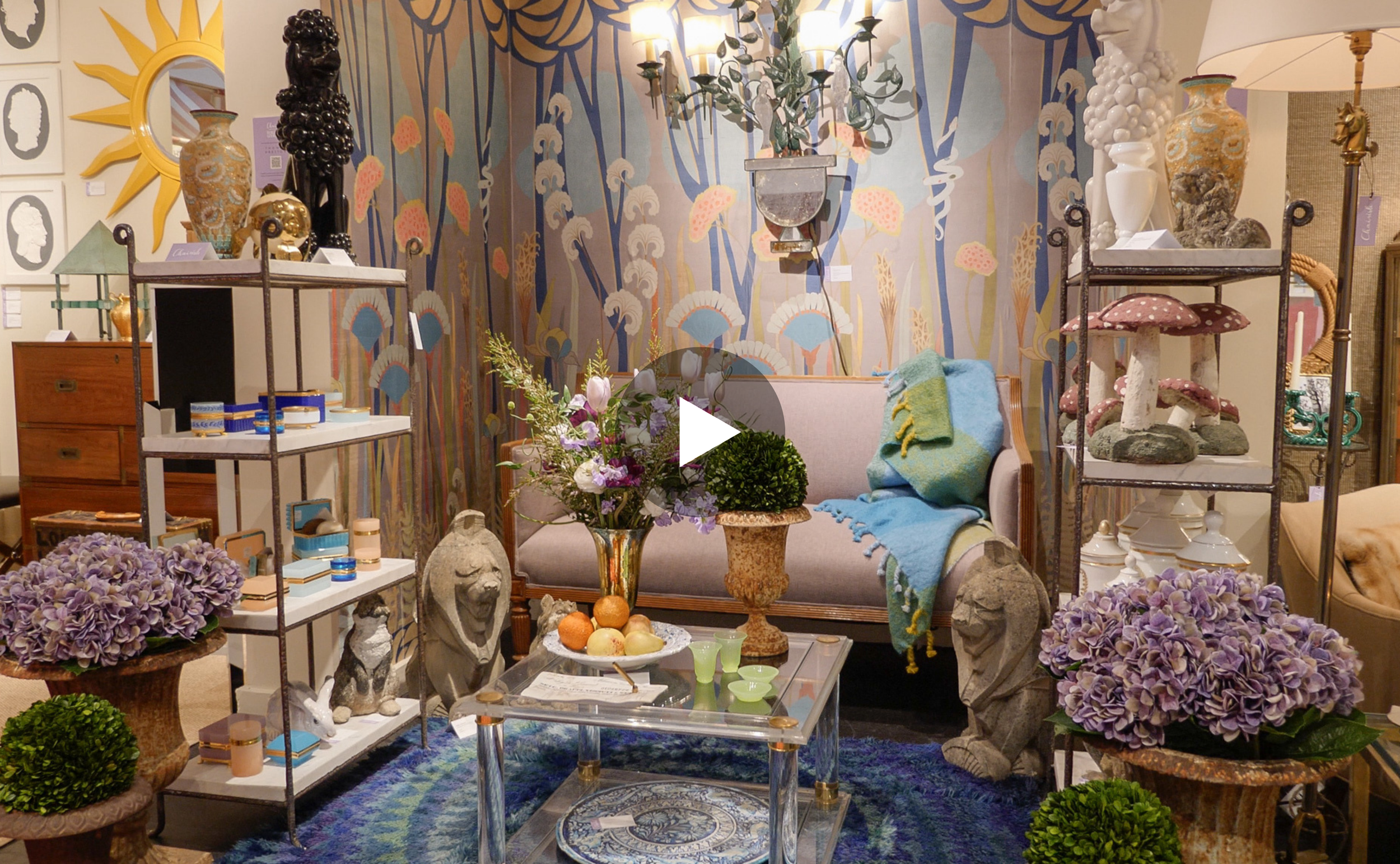 A room decorated with a vintage aesthetic, featuring a floral-patterned wall, a glass coffee table with fruit and paper, purple hydrangeas, sculptures, and various decorative items on shelves and furniture.