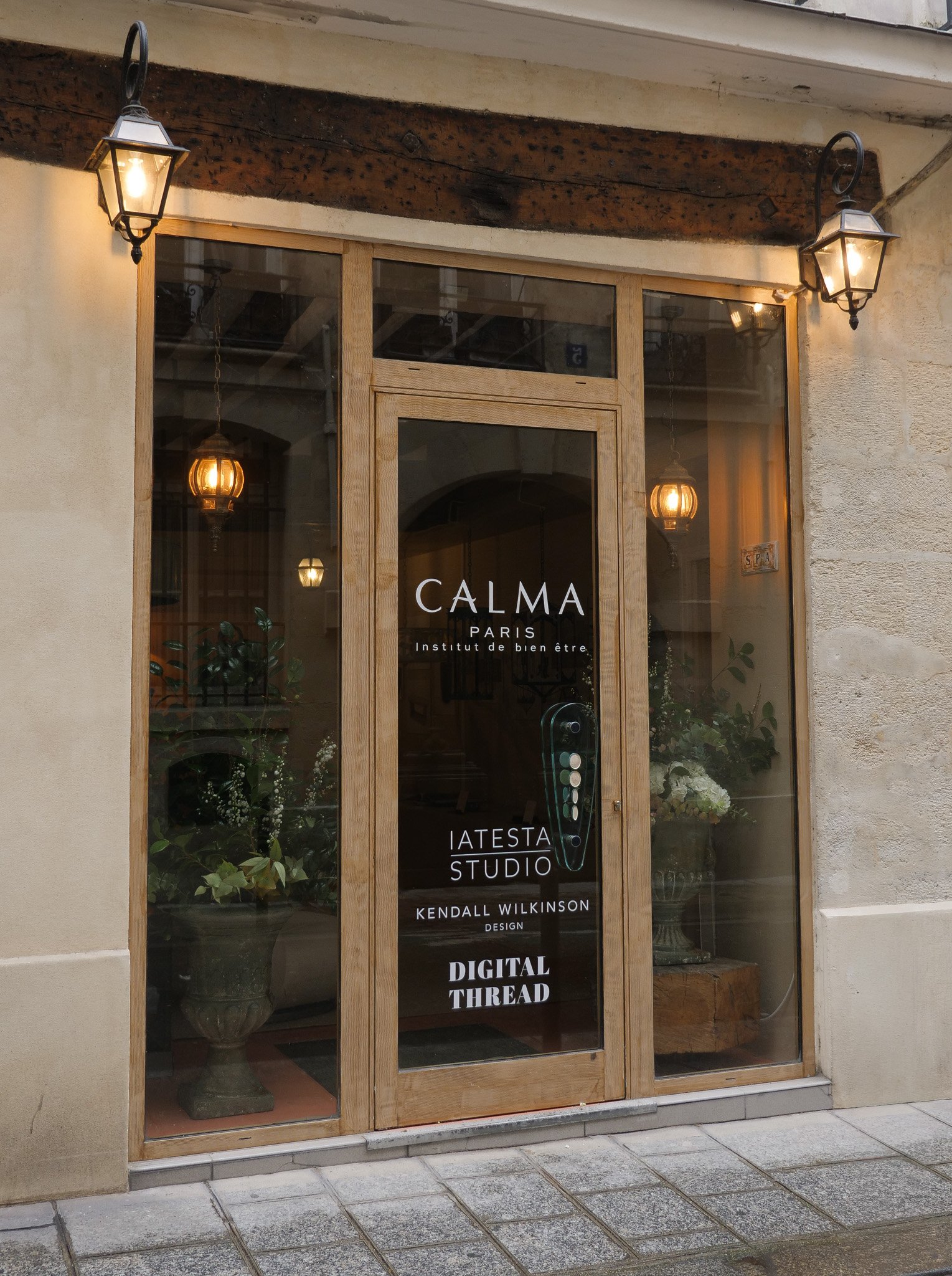 Storefront with glass door and windows, featuring signs for CALMA Paris, Institut de bien être, IATESTA Studio, Kendall Wilkinson Design, and Digital Thread. Inside, there are plants and hanging pendant lights.