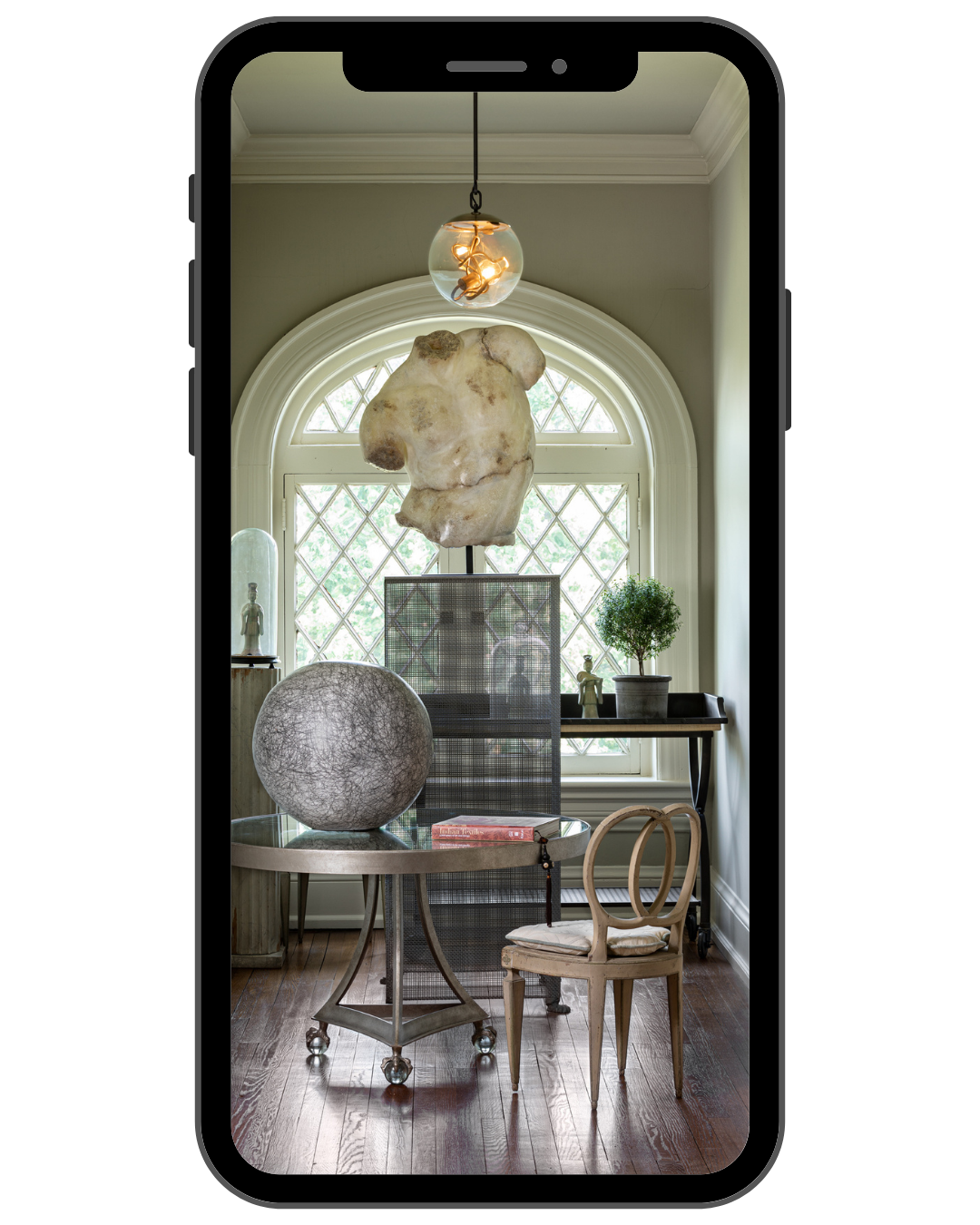 Interior of a room with a large arched window, hanging light fixture, and sculptural decor. There is a table with a large spherical object, a small chair, a potted plant, and a hexagonal decorative screen.