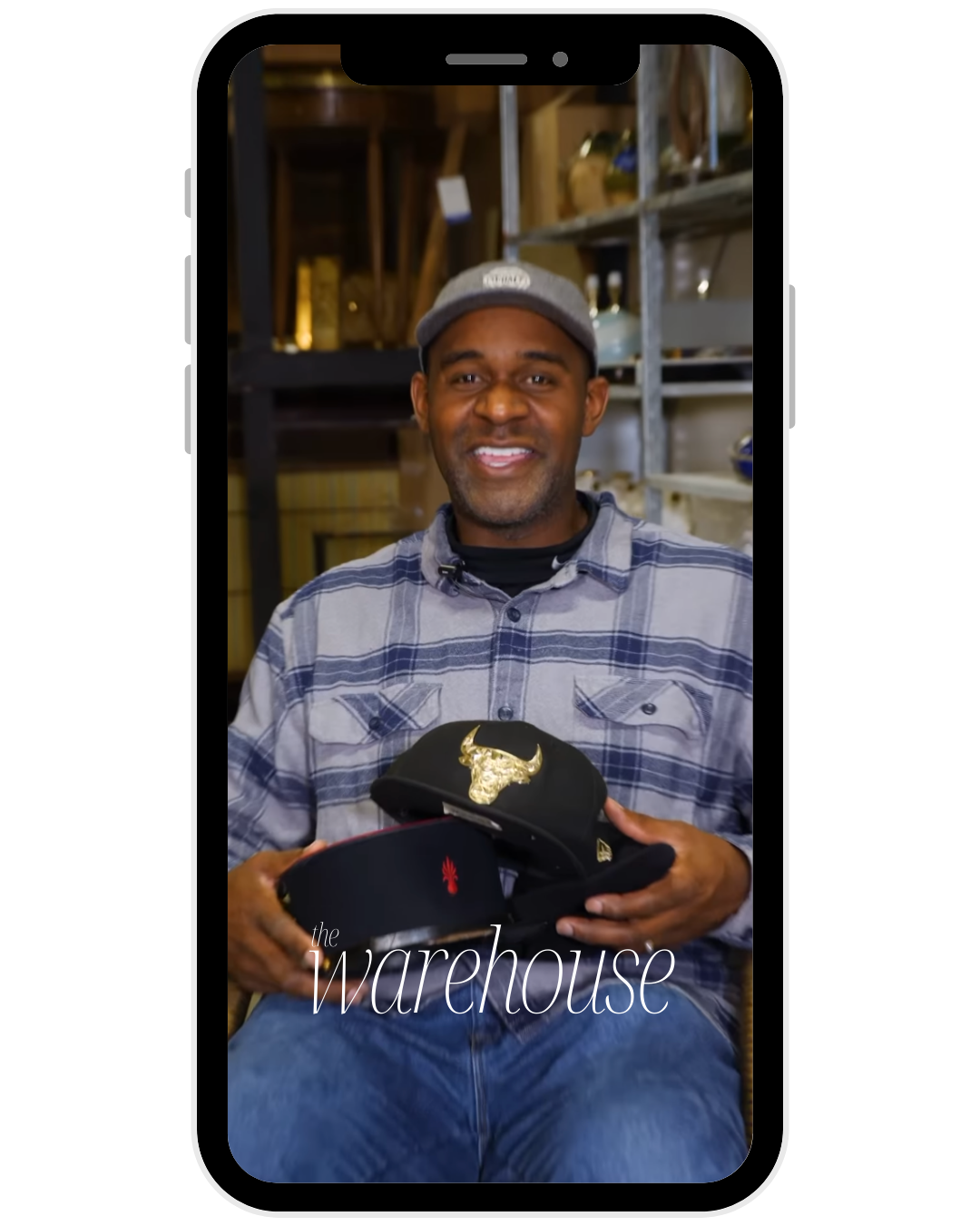 A man smiling and holding a cap with a gold bull logo, sitting in a warehouse setting.