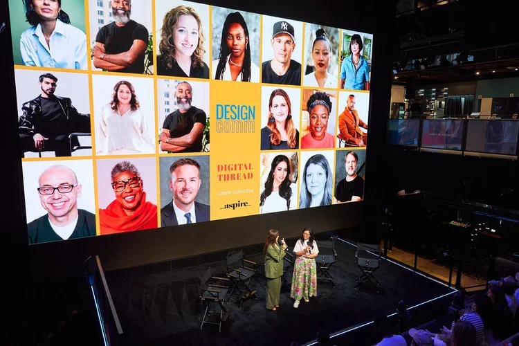 Large screen showing a collage of diverse people, with some photos labeled 'Design Comm' and 'Digital Tribe'. Two women are standing and talking on a stage in front of the screen, while an audience watches. The setting appears to be a conference or presentation event.