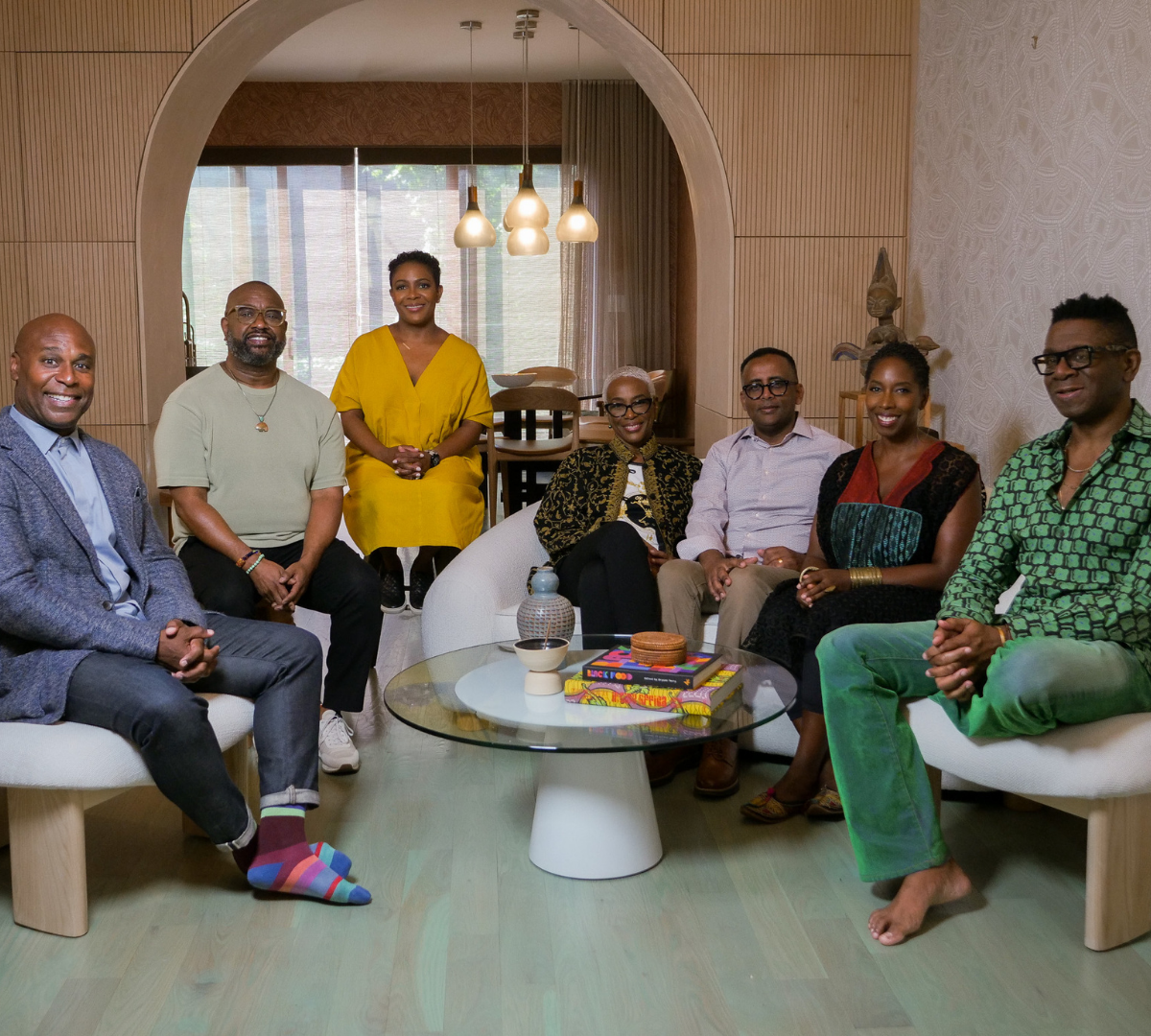 A group of seven diverse adults sitting and standing in a modern living room, smiling for a photo. The room features wooden accents, a round glass coffee table with books and decorative items, and contemporary pendant lights.