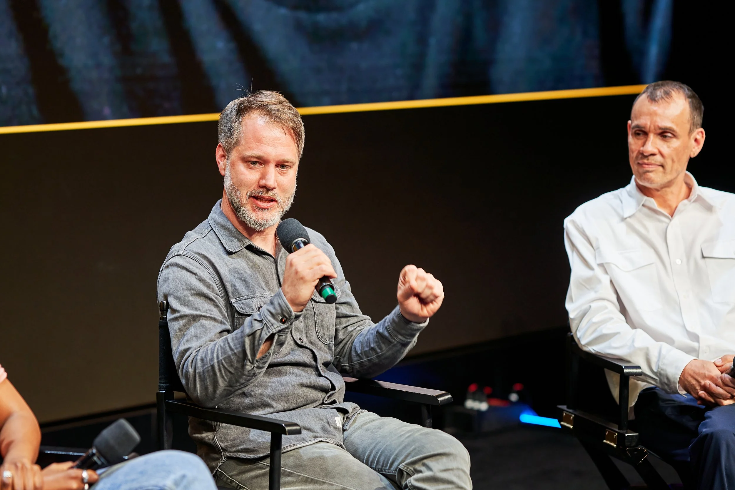 A man with a gray beard and gray hair speaking into a microphone during a panel discussion, sitting on a black chair on a stage with a black background, another man in a white shirt is sitting next to him.