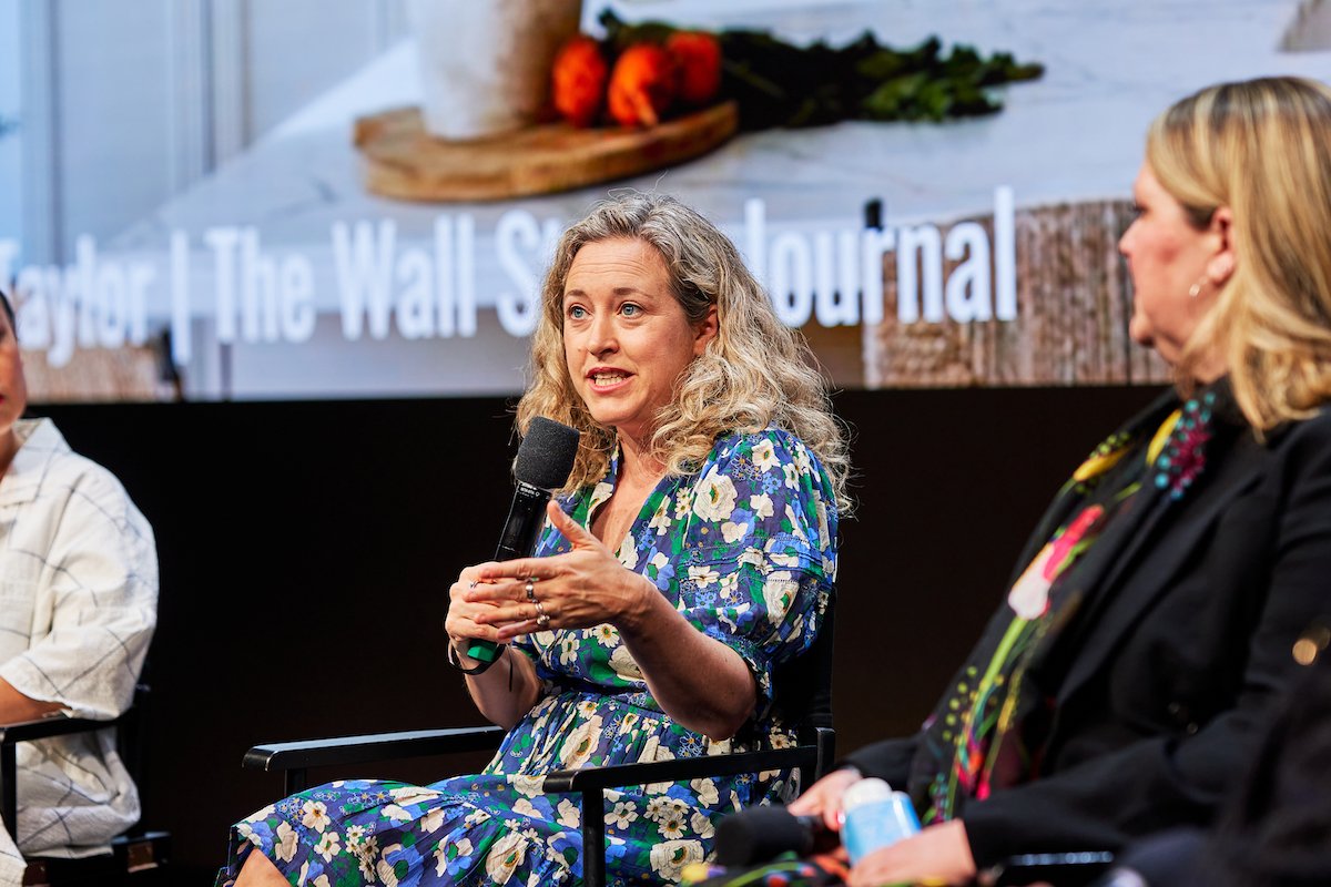Woman with curly blonde hair speaking into a microphone during a panel discussion, with a large screen behind her displaying a logo or title.