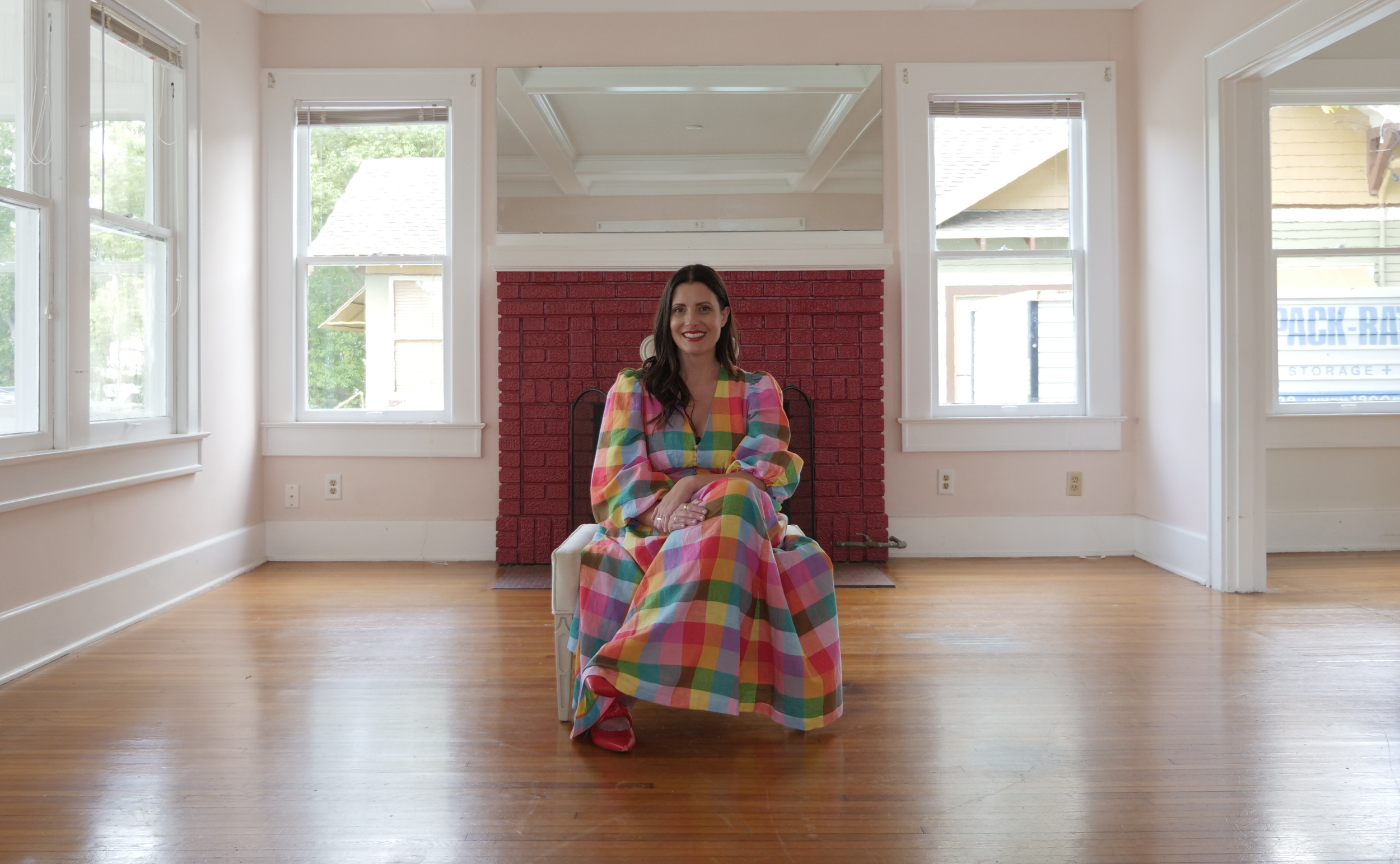 A woman with dark brown hair sitting on a beige chair in front of a red brick fireplace in a bright room with hardwood flooring, white trim, and large windows.