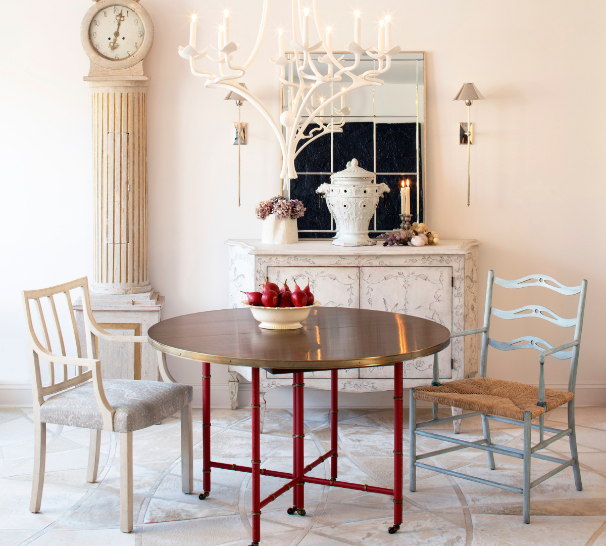 A dining room with a round wooden table, two different chairs, a sideboard with decorative items, a mirror, a tall antique clock, and a chandelier above.