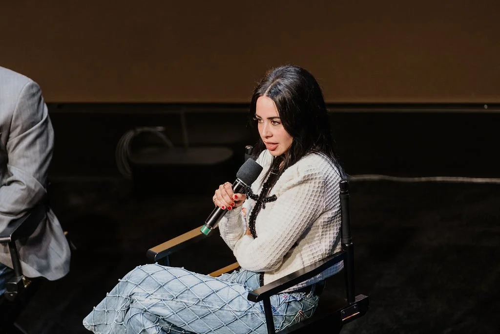 Woman with black hair sitting on a chair, holding a microphone, participating in a panel or discussion.