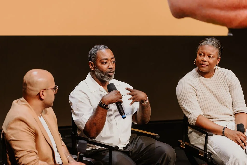 Three people sitting on chairs participating in a panel discussion or interview, with a man in the middle speaking into a microphone.