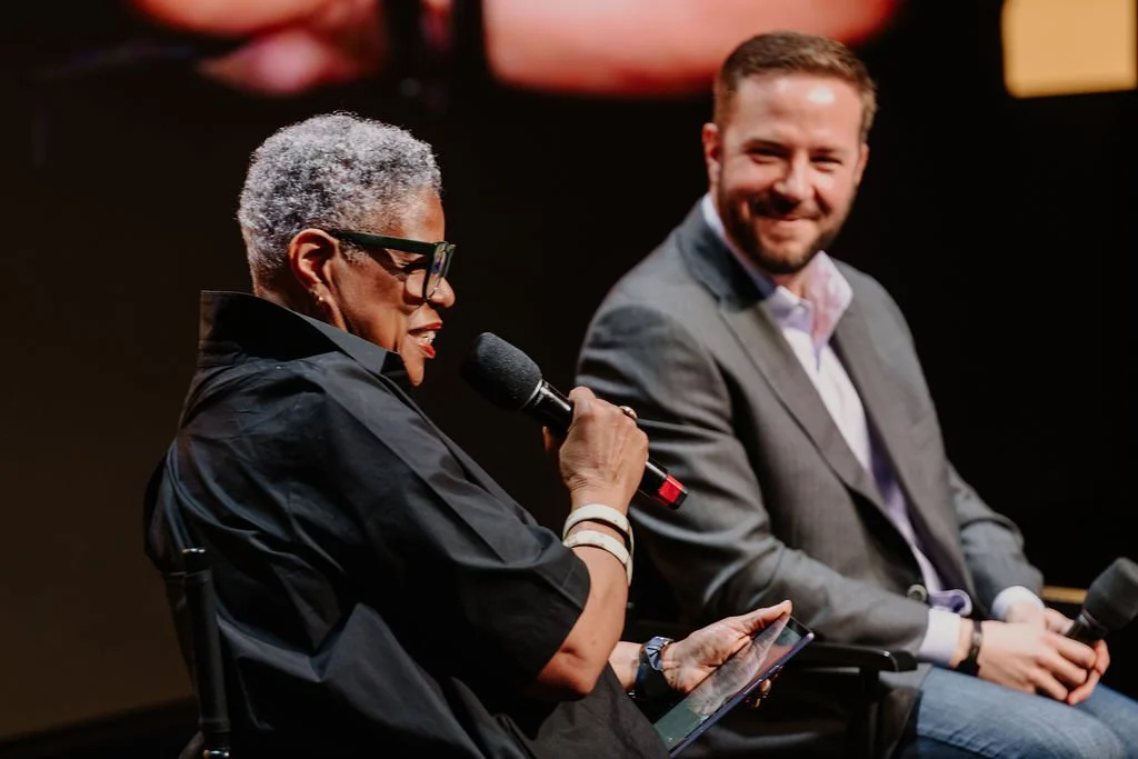 A woman with short gray hair, glasses, and earrings is speaking into a microphone while holding a tablet, seated next to a man with short brown hair and a beard, wearing a gray blazer and white shirt, who is smiling and holding a microphone.