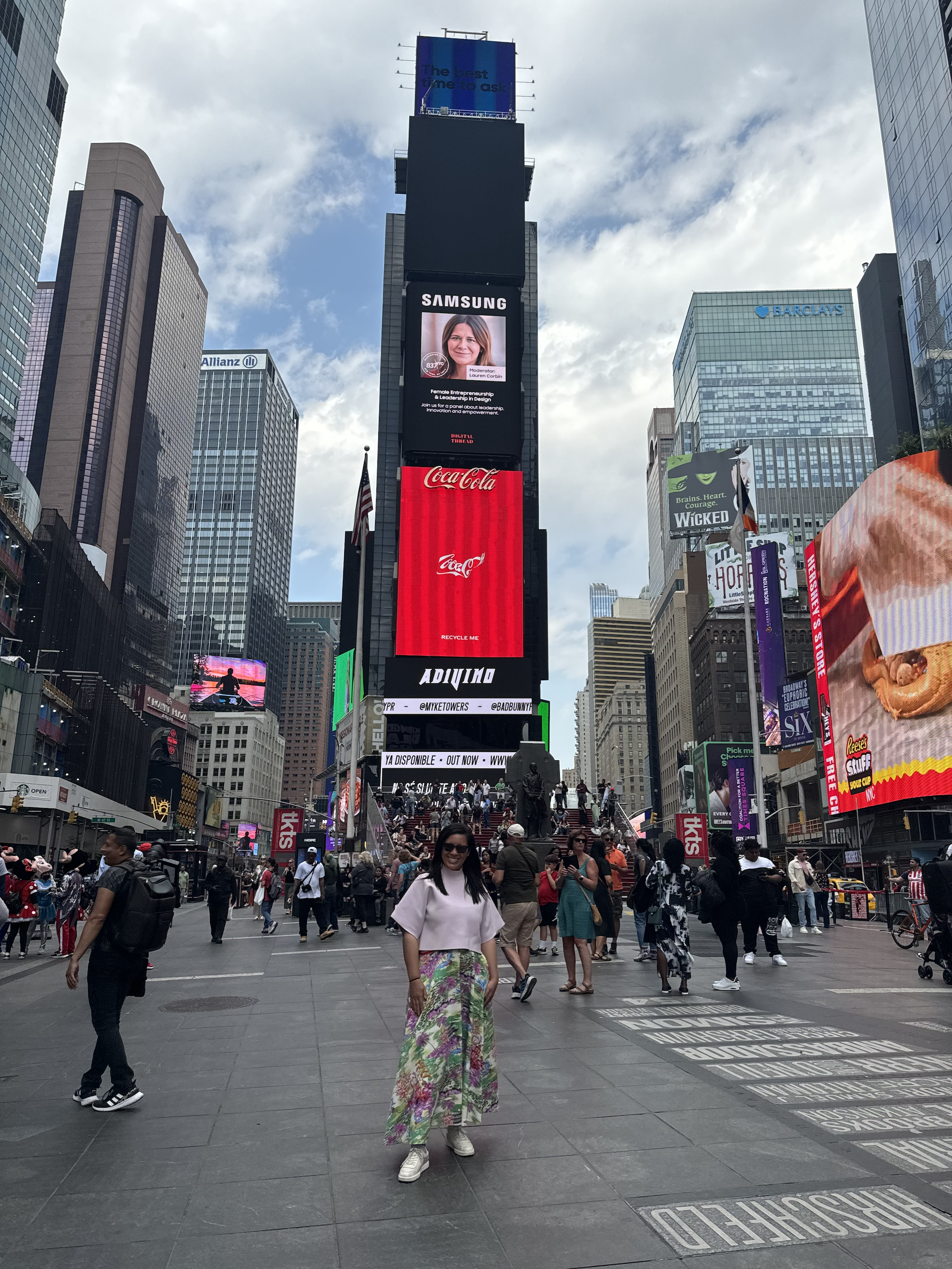 Woman standing in Times Square, New York City, with large digital billboards and tall buildings around her, people walking and taking photos, and the sky partly cloudy.