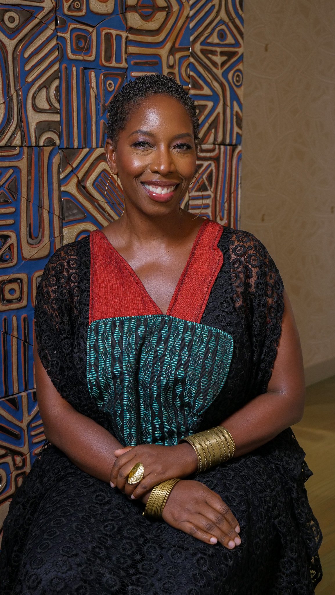 A woman with short, curly hair wearing a black lace dress with red and teal accents, sitting with her hands on her lap, smiling in front of a decorative wooden wall.