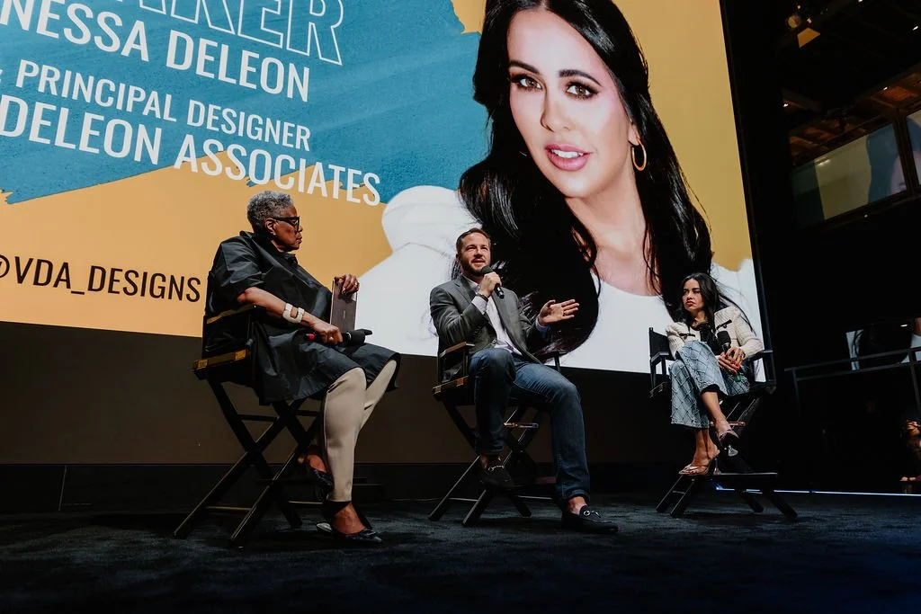 Panel discussion on stage with three people sitting in director's chairs in front of a large screen displaying an image of a woman and text. The woman on the screen has dark hair, hoop earrings, and is smiling.