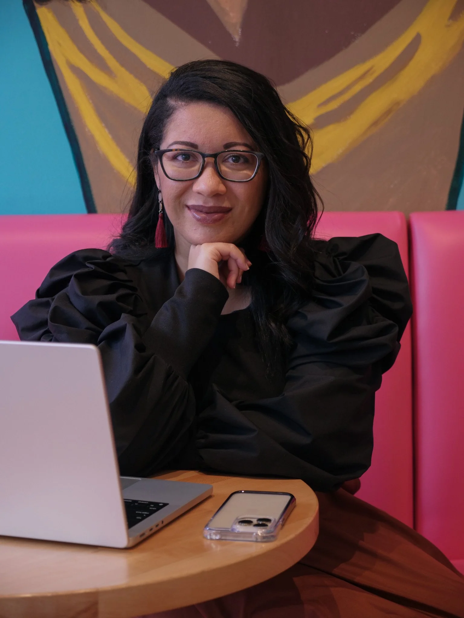 A woman with glasses, black hair, and wearing a black top, sitting at a table in a cafe or restaurant with a laptop and a smartphone, with pink seating and abstract art on the wall behind her.