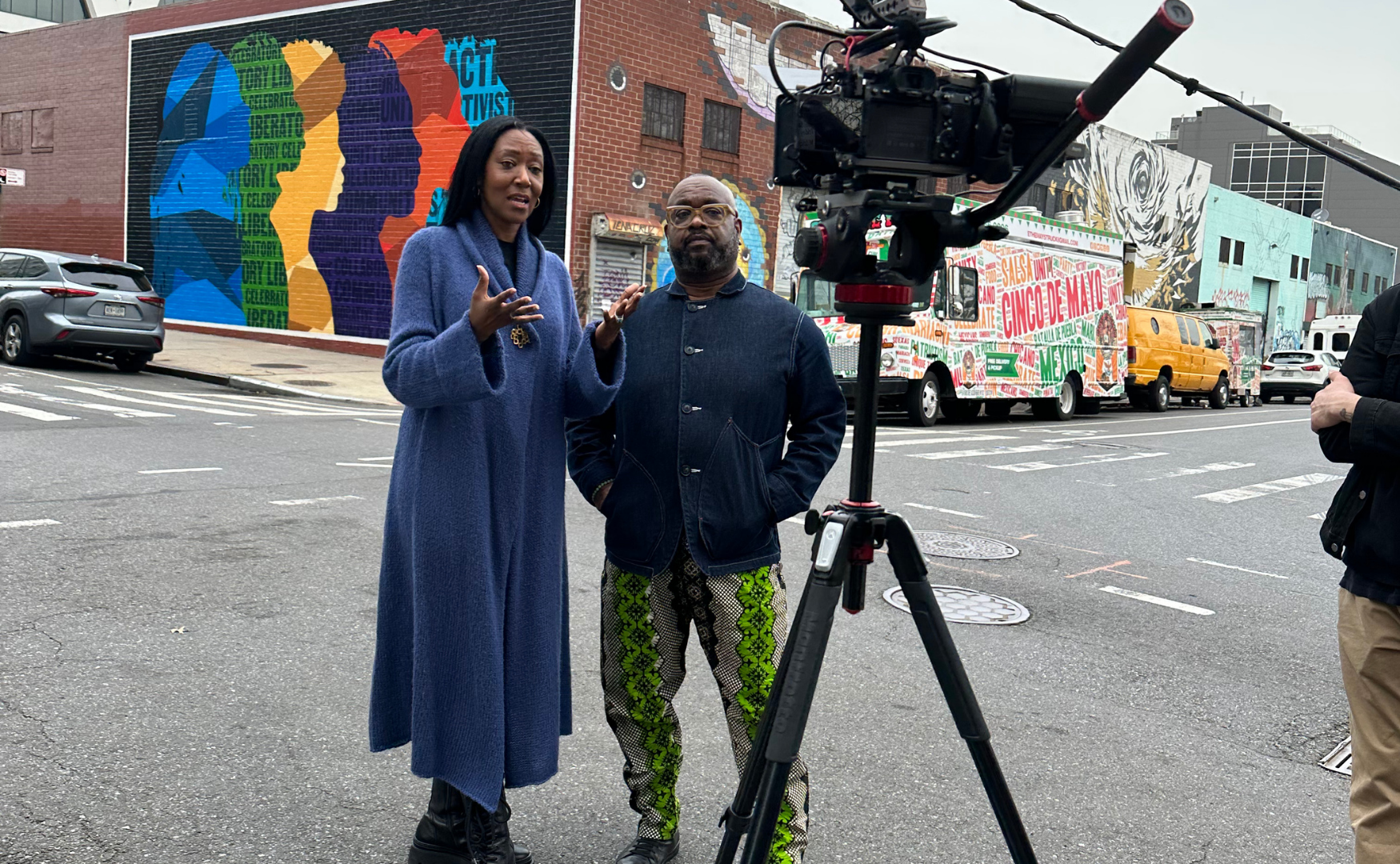 Two people standing outdoors in front of a camera on a tripod, engaged in conversation. Behind them are colorful murals and parked vehicles.