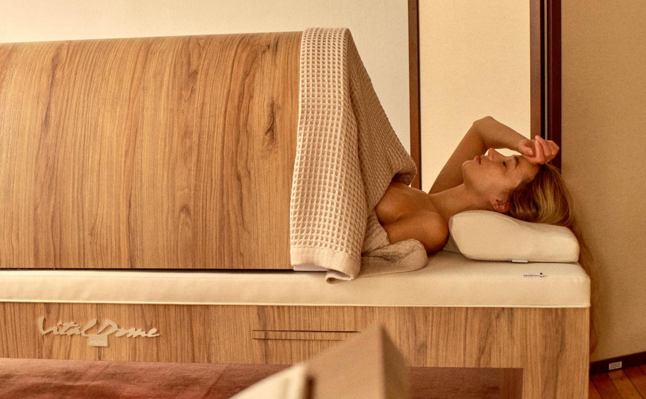A woman lying on her side on a hotel bed with a beige headboard, resting her head on a pillow, with her arm bent above her head, in a relaxed pose.