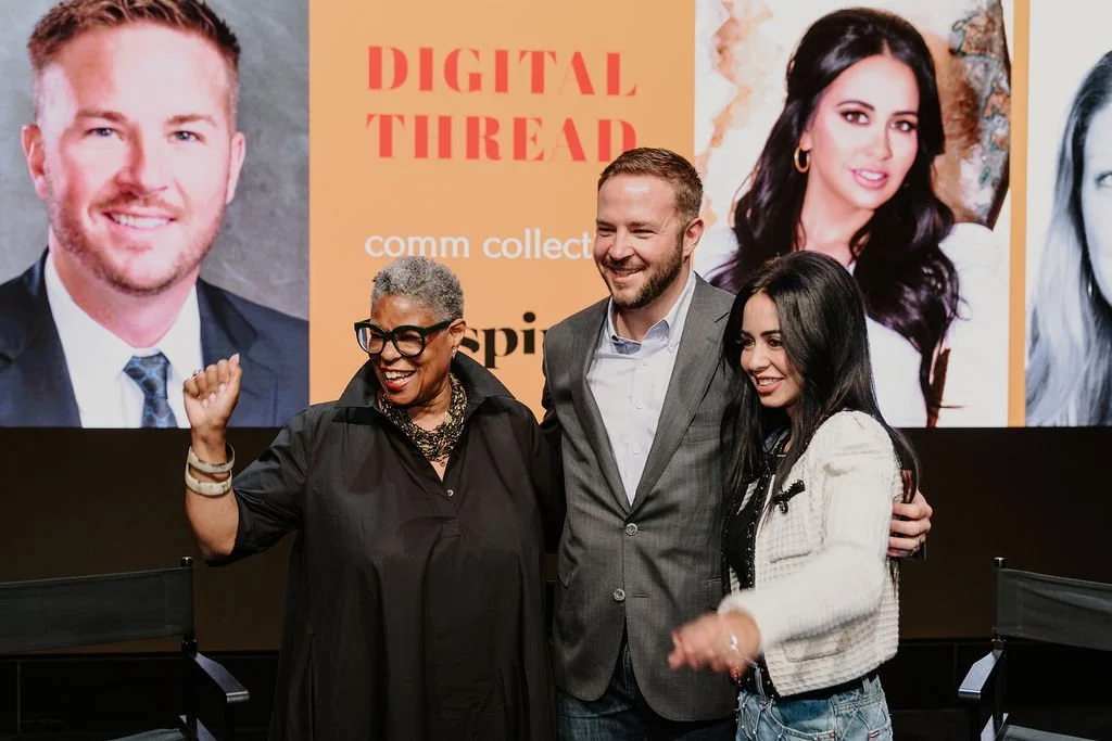 Three people standing together on stage, smiling and posing for the camera in front of a large screen showing headshots of other individuals. The background has an orange section with text reading 'Digital Thread' and other words partially visible.