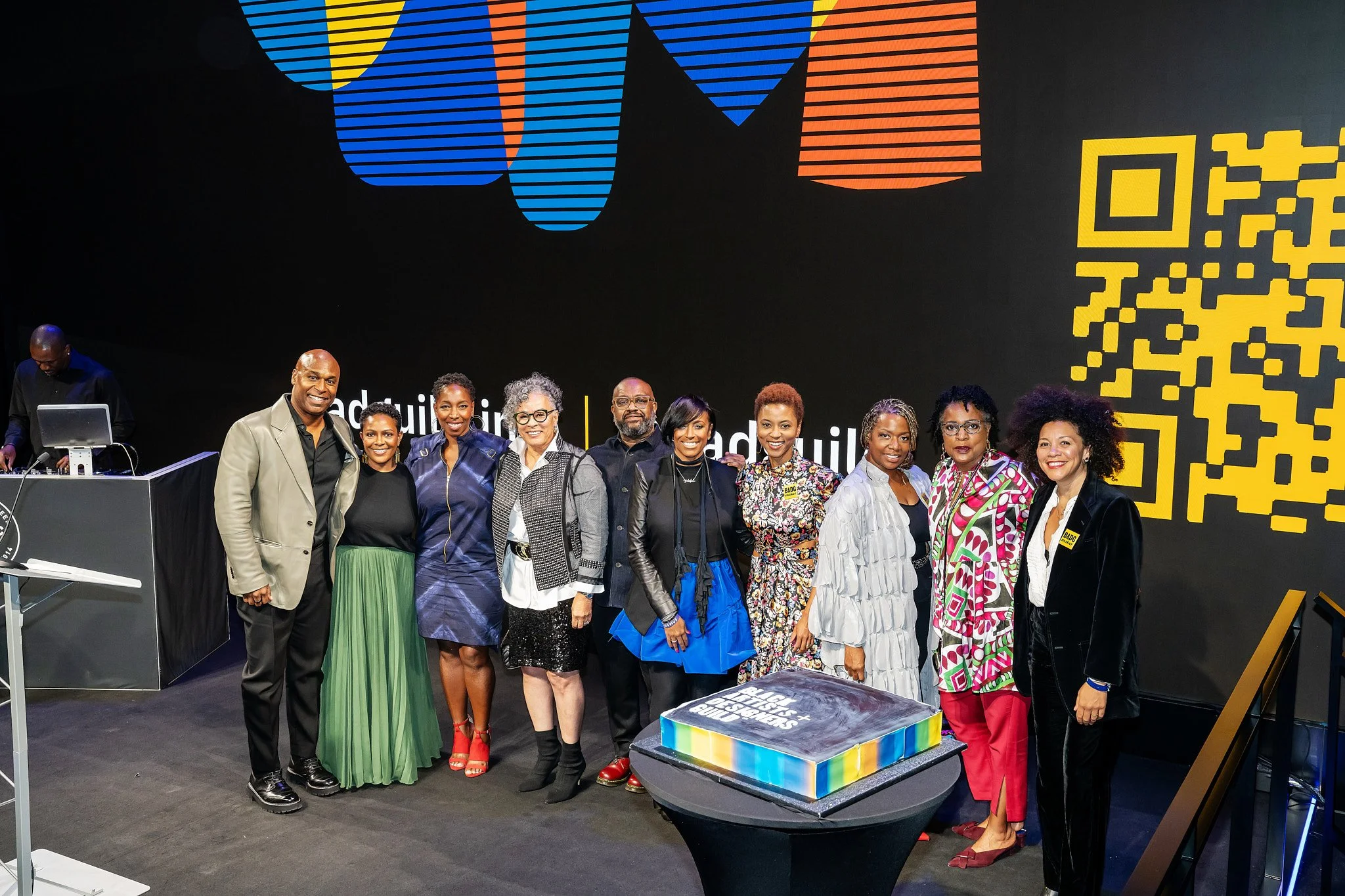 Group of diverse people standing on stage, celebrating at an event, with a large black background featuring colorful shapes, a QR code, and a cake on a table in front.