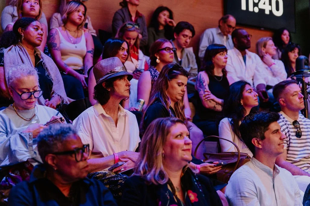 A diverse group of people sitting in a theater or auditorium, attentively watching a presentation or event. Some are using phones, while others are focused on the stage. The background shows a digital clock displaying 1:40.