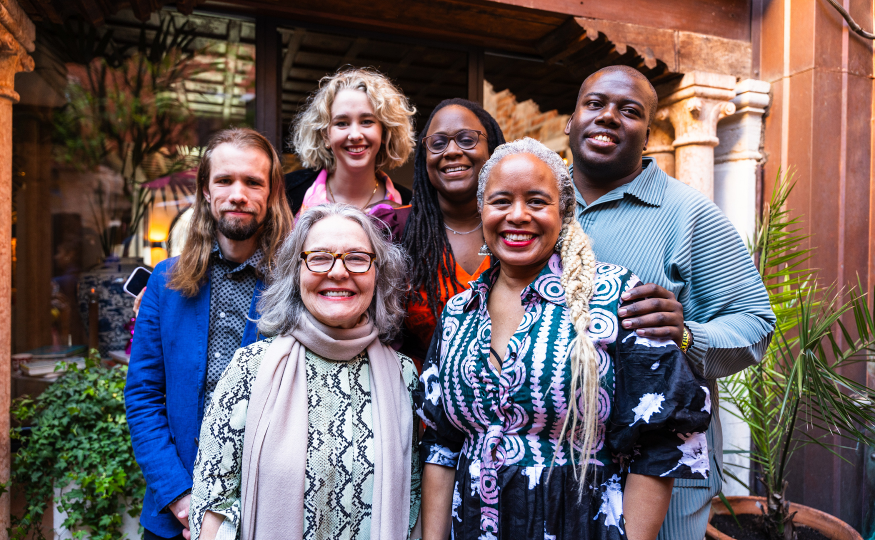 Group of seven diverse people smiling and posing together indoors with plants and architectural decor in background.