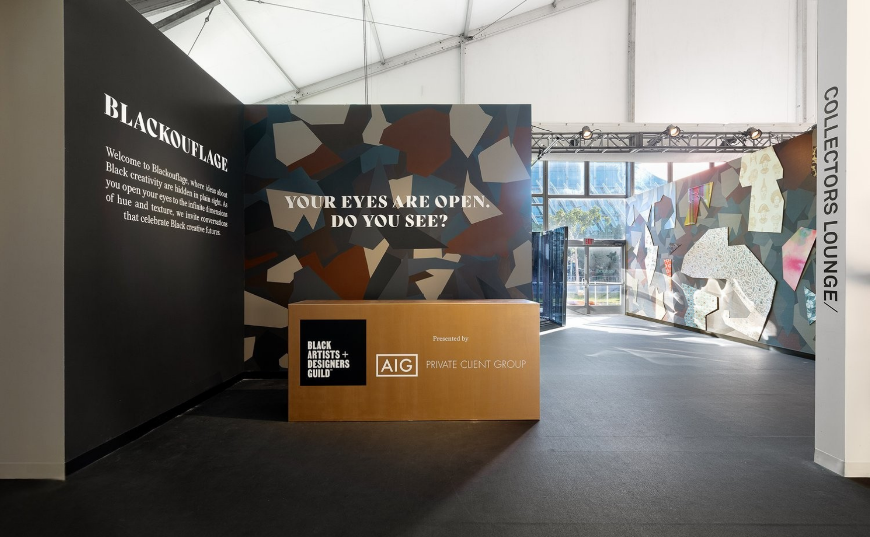 An art exhibition entrance with a black wall on the left, a colorful geometric mural, and a reception desk in the center. Text on the wall reads "YOUR EYES ARE OPEN. DO YOU SEE?" and the event is titled "BLACKOUFLAGE" celebrating Black creativity, pr
