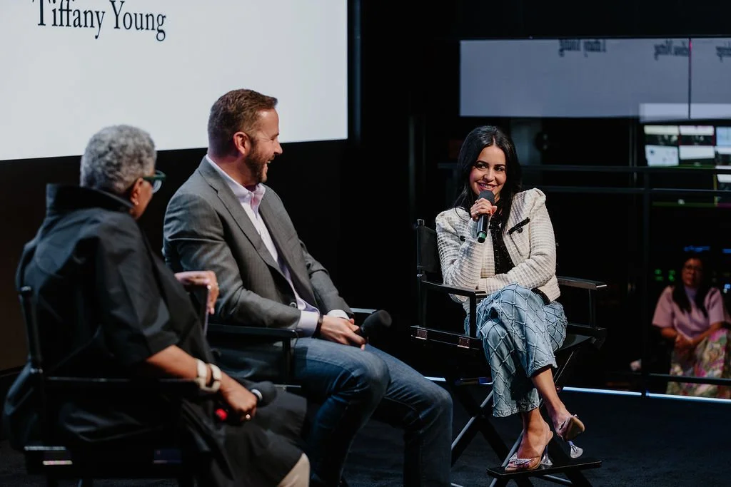 Three people sitting on a stage engaging in a discussion, with a woman holding a microphone and smiling.