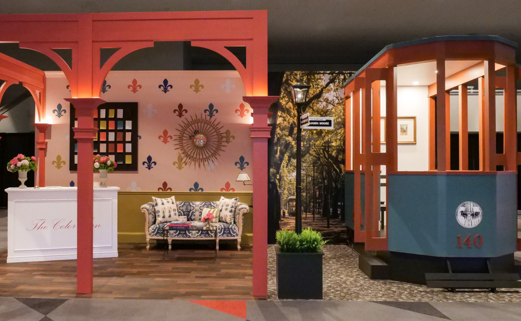 Indoor display of a salon or boutique with a vibrant pink and blue color scheme, featuring a sitting area with a floral sofa, decorative wallpaper with fleur-de-lis patterns, an ornate clock, and a small stage or lift with a framed photo on the wall.