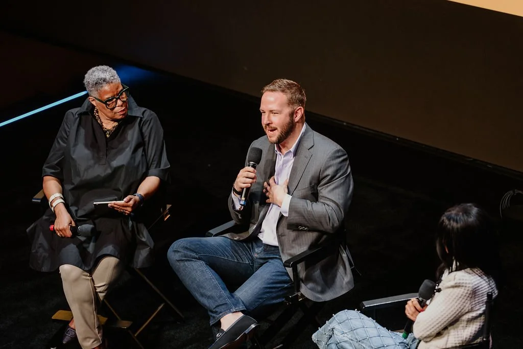 A panel discussion with three people on stage, two women and one man. The woman on the left has short gray hair, glasses, and is wearing a black outfit. The man in the middle has short hair, a beard, wearing a gray blazer, white shirt, and jeans, and