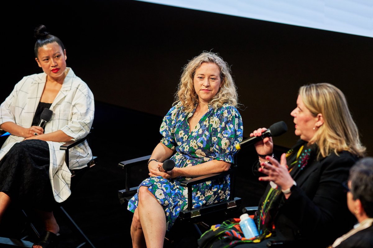 Three women sitting on a panel in front of a black background, two holding microphones and one listening attentively.