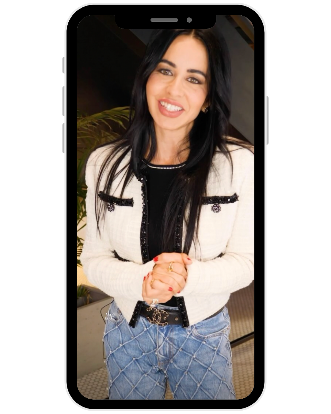 A woman with long black hair, smiling, wearing a cream-colored jacket with black trim, a black top, and blue jeans with a diamond pattern, standing indoors.