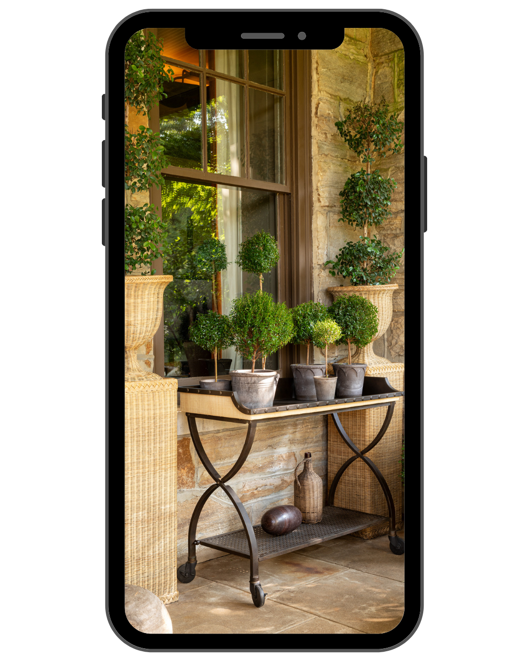 Indoor patio with potted plants on a rolling metal cart, wicker chairs, a large window, and decorative jars against a stone wall.