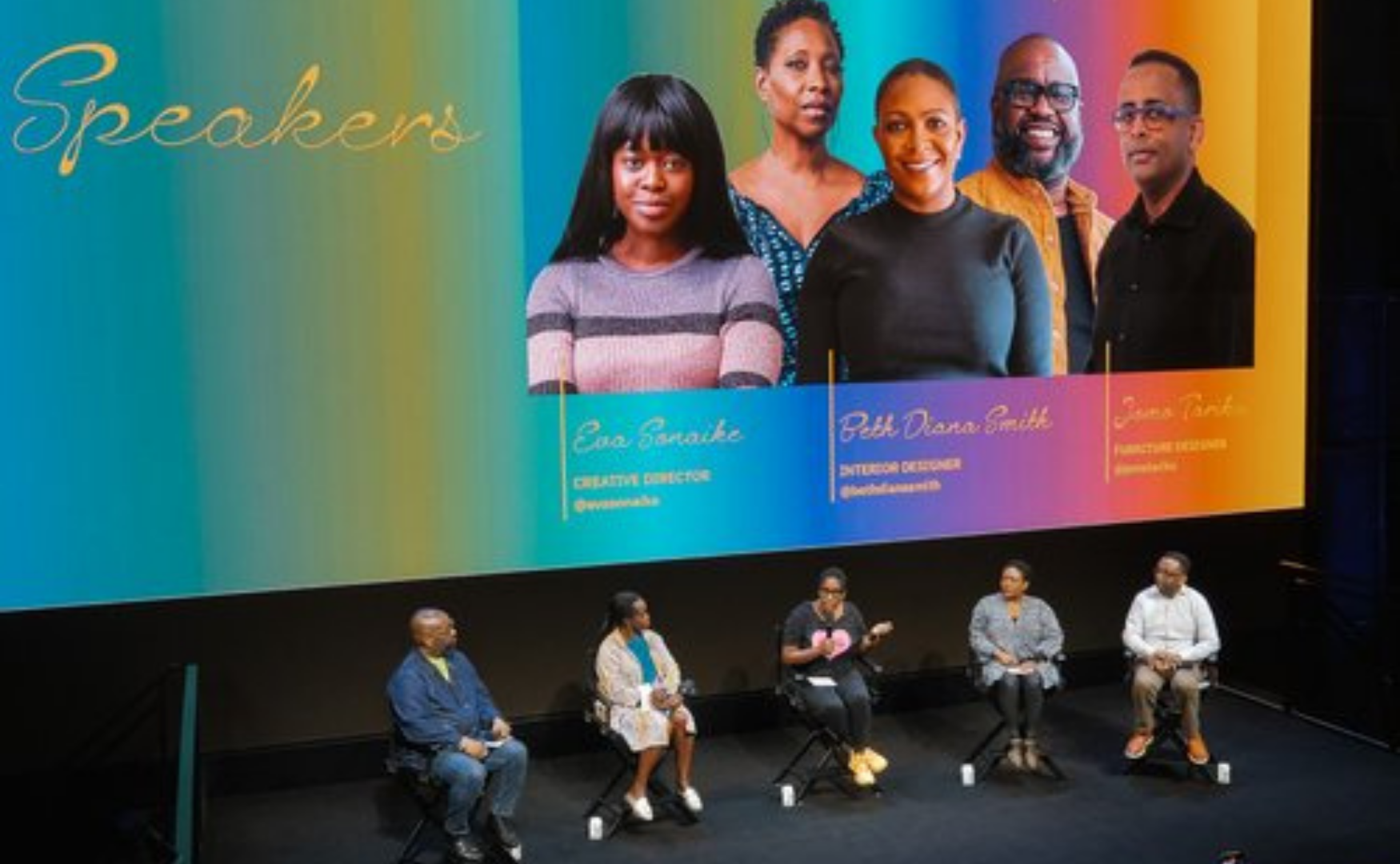 Panel discussion on stage with five people seated in front of a large screen displaying profiles of three women, with colorful background.