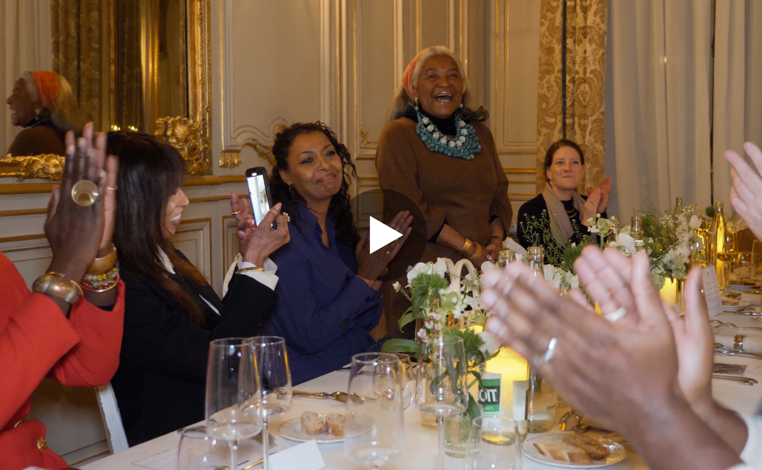 Women sitting at a banquet table clapping and smiling, with one woman standing and speaking, in an elegant decorated room.