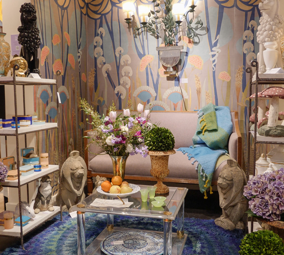 An interior room decorated with vintage and modern decor. Features a beige sofa with a colorful throw blanket, a glass coffee table with a floral arrangement, and various sculptures and ceramics on shelves. The background wall has a nature-inspired mural with plants and dandelions.